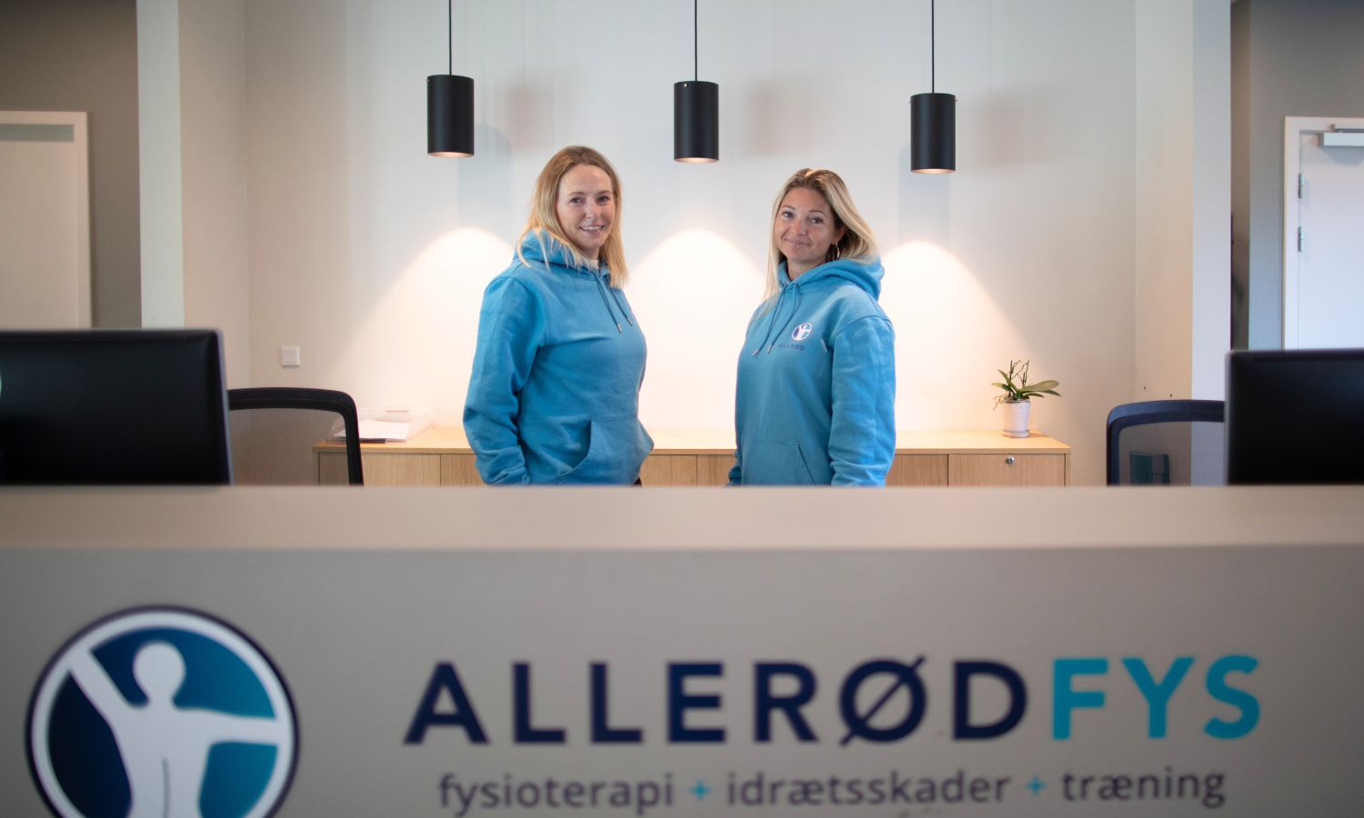 To medarbejdere smiler i receptionen hos Allerød Fys.